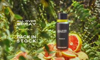 All Over Spray bottle with magnolia scent on a background of green leaves and sliced fruits.