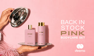 A pink bodycare set by Pini on a pink background, held by hands on a silver tray with a reflective bowl. The set includes a pink bottle and a pink jar with gold caps, alongside a large text overlay reading 'BACK IN STOCK PINK BODYCARE SET' and the 'deema' logo.